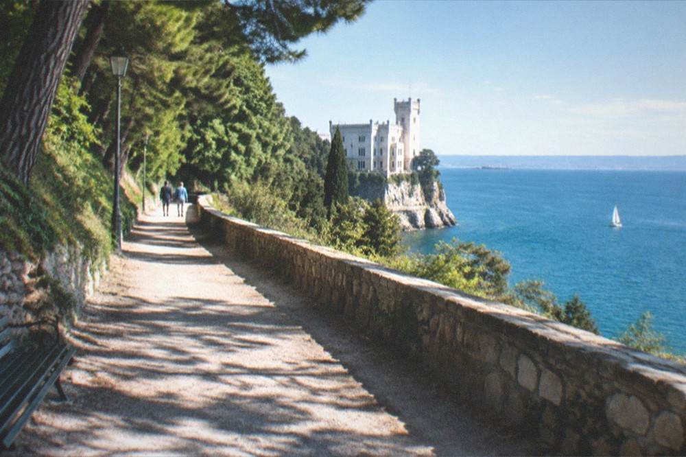 A wooded park path above the sea leading towards a white cliff-top castle