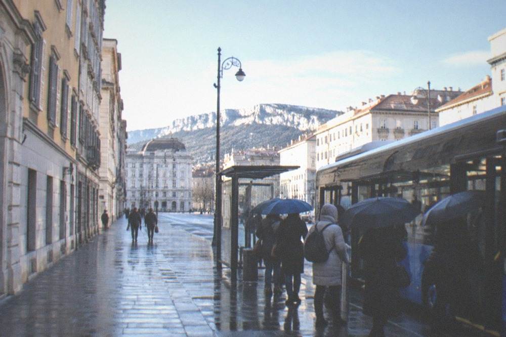 A city bus at a wet stop in Trieste with passengers boarding under umbrellas