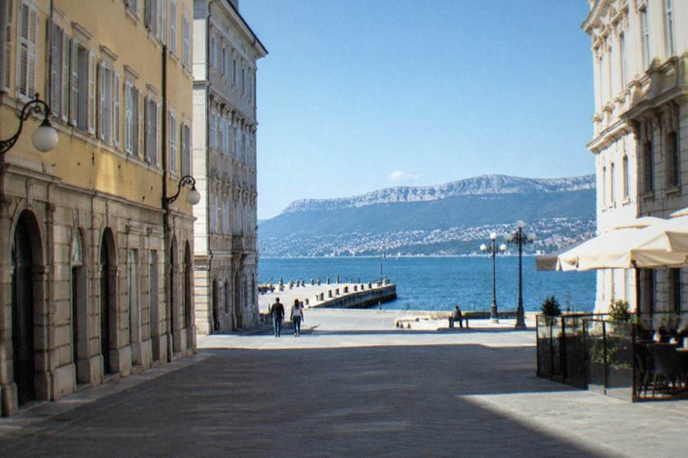 A stone pier reaching into a calm blue gulf with hills on the far shore