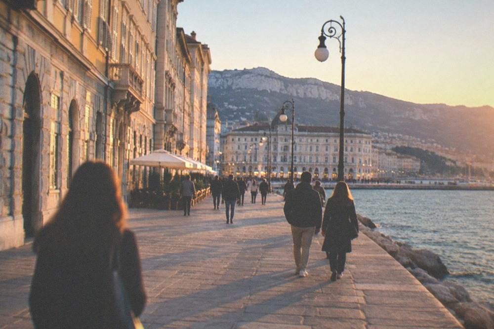 People walking the seafront at dusk with the lights of the piazza behind
