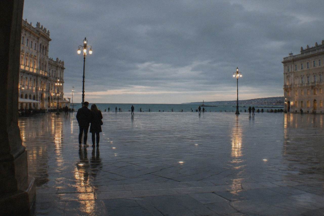 A couple standing on Trieste's seafront at dusk with the lights of Piazza Unità d'Italia reflected on wet paving