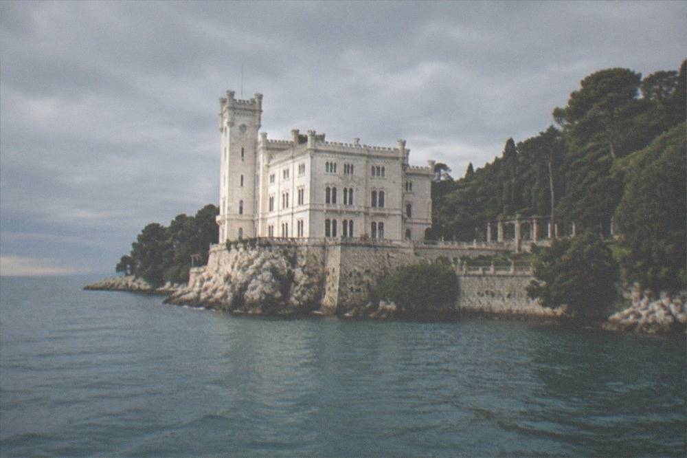 Miramare Castle seen from the sea with its white facade above the water and the park trees on either side
