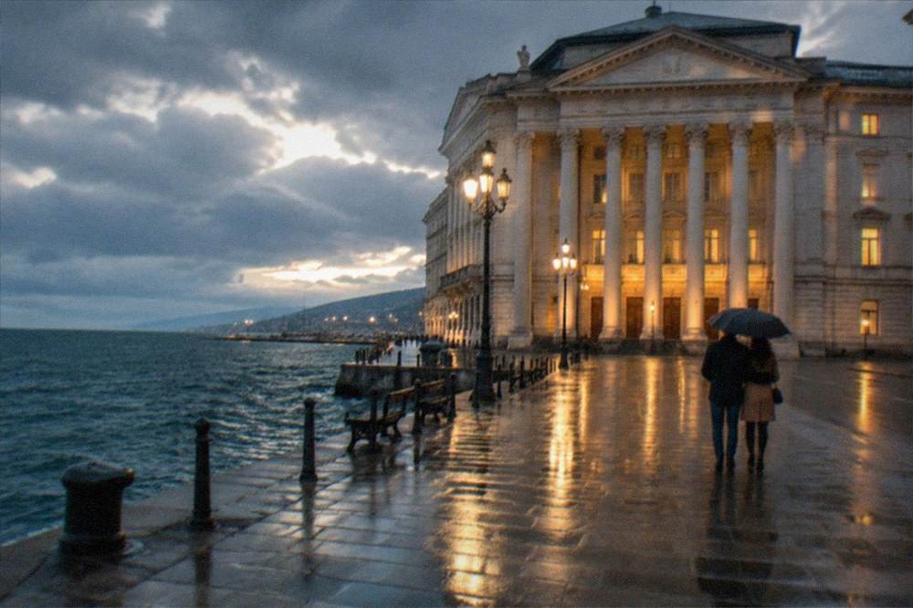 A couple sharing an umbrella on a rainy evening in front of the lit facade of the Teatro Verdi in Trieste