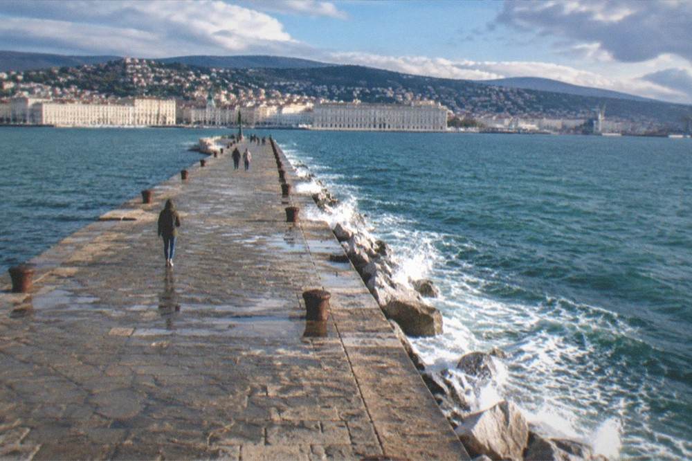 The Trieste seafront promenade under a clearing sky, white spray on the rocks of the Molo Audace pier