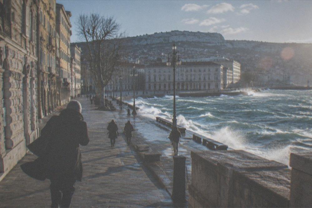 A walker leaning into strong wind on the Trieste seafront with whitecaps on the gulf